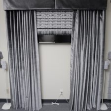 Draperies under an inverted pleat valance and over a roman shade, all made out of a grey velvet fabric and all concealing a TV.
