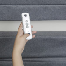 A woman's hand holding a small remote with three buttons on it in front of two shades.