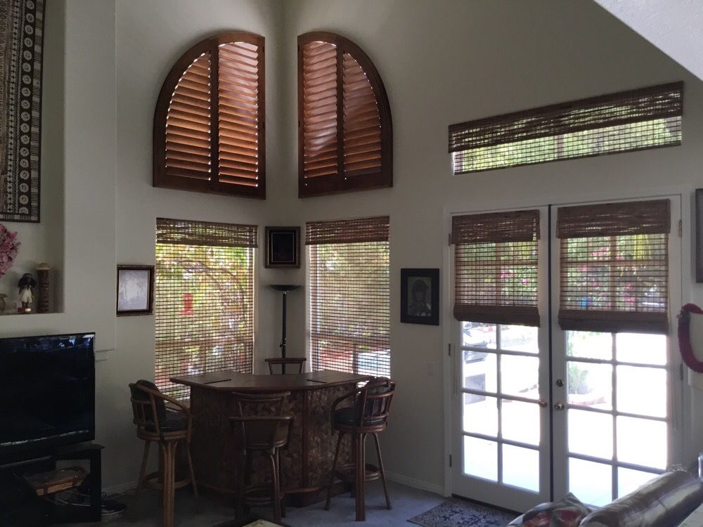 Corner of a living room with shutters covering quarter circle windows above four windows covered in woven wood window shades.