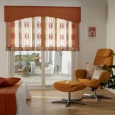 Transparent floral orange curtains half up on sliding glass door in the corner of the room with a lounge chair facing outside.