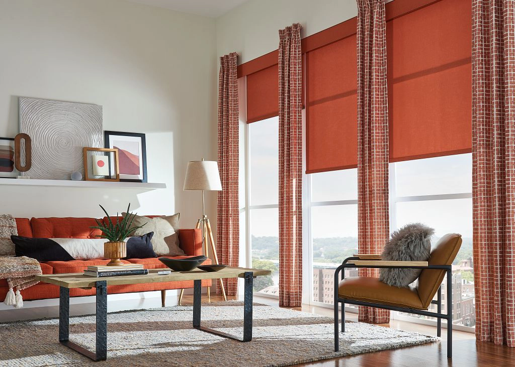 a bright orange-red living room color is visible on the couch, the stationary drapery panels and the solar shades in the large windows. its color is balanced with white walls, a white rug, and light wood flooring