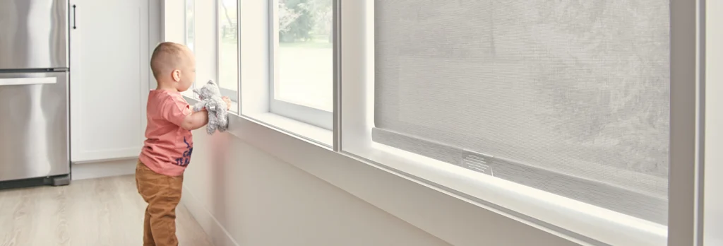 a toddler stands by a kitchen window holding a toy - cordless window treatments are necessary for child safety
