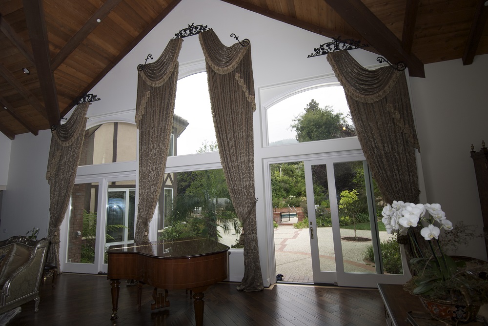 Three top treatment drapery swags elegantly hanging over three large windows in a high ceiling living room.