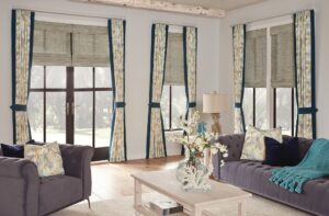 Living room with three large windows. Pleated drapes are pulled back and roller shades are drawn halfway up the windows